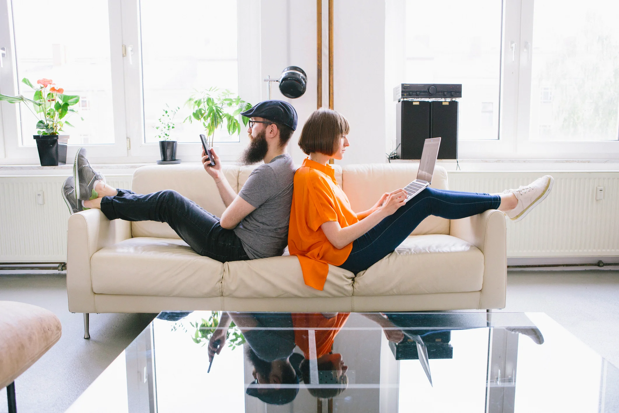 Junges Paar sitzt Rücken an Rücken auf Sofa, nutzt Laptop und Smartphone – Symbolbild für digitale Antragstellung bei einer Mietkautionsversicherung.