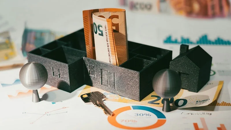 Symbolbild mit Hausmodellen, Euro-Banknoten, Schlüssel und Diagrammen – Darstellung finanzieller Aspekte rund um Mietverhältnisse und die Rolle der Mietkautionsversicherung.