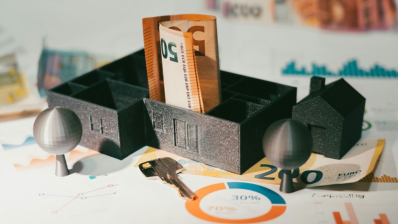 Symbolbild mit Hausmodellen, Euro-Banknoten, Schlüssel und Diagrammen – Darstellung finanzieller Aspekte rund um Mietverhältnisse und die Rolle der Mietkautionsversicherung.