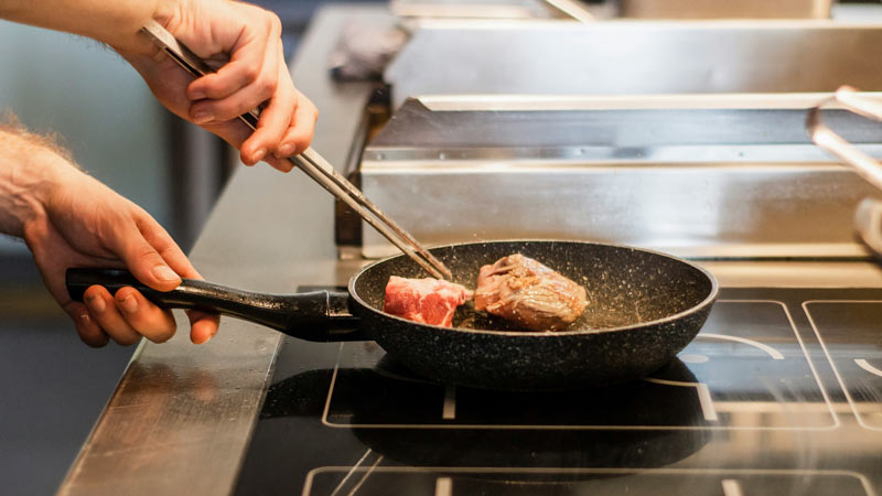 Hand brät Fleisch in einer Pfanne auf einem modernen Induktionsherd – Symbolbild für Gebrauch der Mietwohnung, Küchennutzung und mögliche Schäden im Kontext der Mietkautionsversicherung.