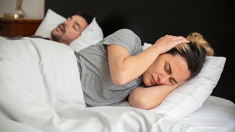 Frau hält sich im Bett genervt die Ohren zu, während ein Mann schnarcht – Symbolbild für Konflikte im Mietverhältnis durch Lärmbelästigung und mögliche Auswirkungen auf die Mietkautionsversicherung.