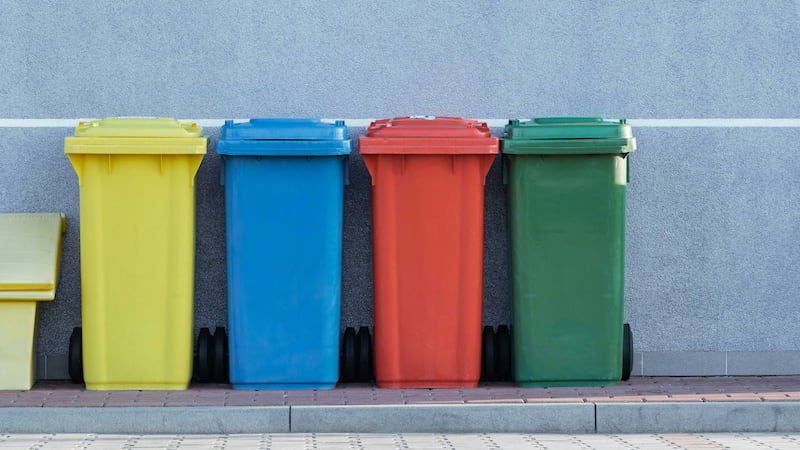 Vier Mülltonnen in Gelb, Blau, Rot und Grün vor grauer Wand – Symbolbild für Mülltrennung und ordnungsgemäße Nutzung gemeinschaftlicher Flächen im Mietverhältnis im Zusammenhang mit einer Mietkautionsversicherung.
