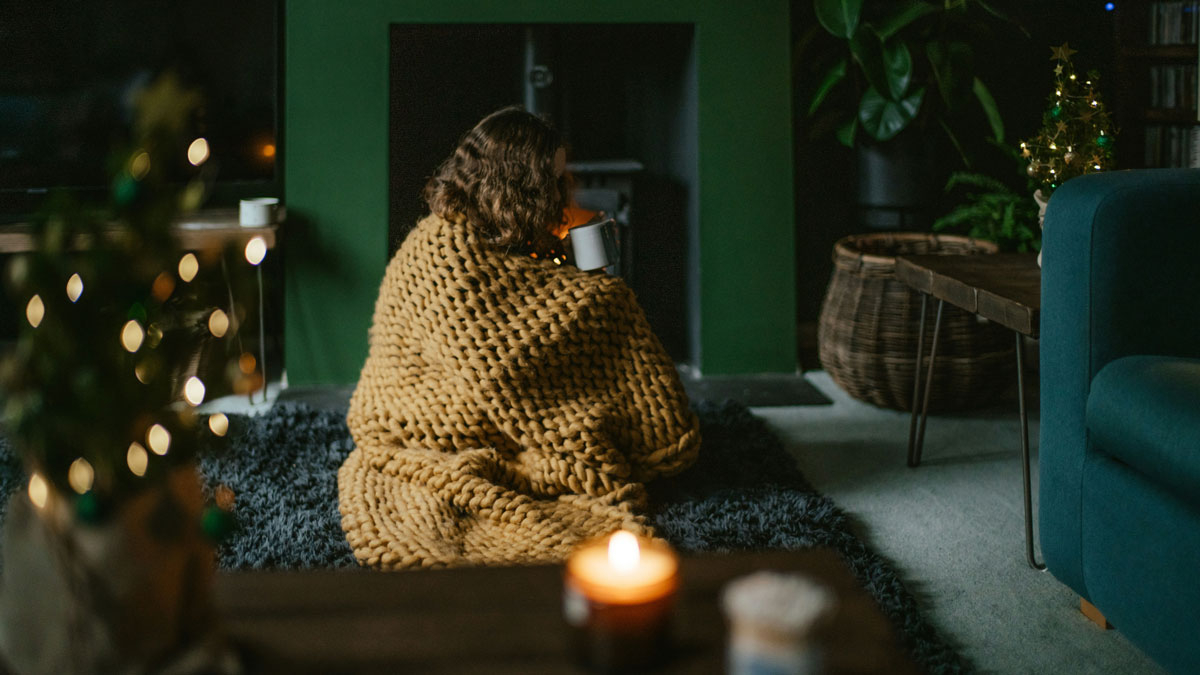Person sitzt gemütlich eingehüllt in eine gestrickte Decke vor dem Kamin und trinkt Tee – Symbol für Geborgenheit und Sicherheit im eigenen Zuhause durch Mietkautionsversicherung.