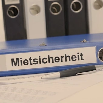 Blauer Aktenordner mit der Aufschrift „Mietsicherheit“, daneben ein Kugelschreiber und Dokumente – Symbolbild für Verwaltung und Absicherung von Mietverhältnissen durch eine Mietkautionsversicherung.