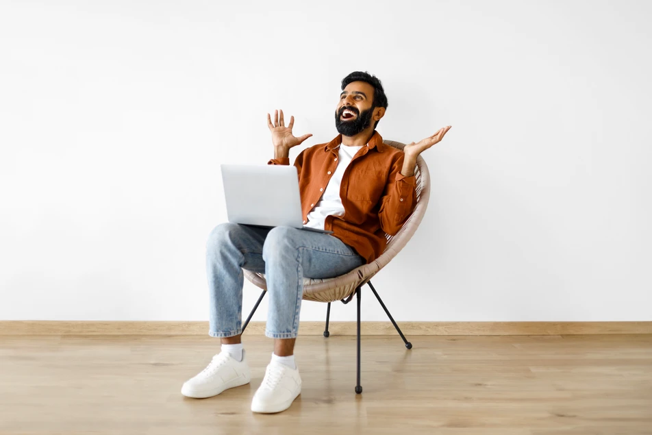 Glücklicher Mann sitzt entspannt mit Laptop auf einem Stuhl und jubelt – Symbolbild für die einfache und erfolgreiche Online-Beantragung einer Mietkautionsversicherung.