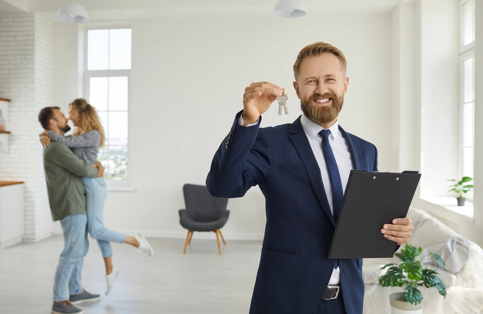 Vermieter hält Wohnungsschlüssel und Unterlagen, im Hintergrund Mieter bei der Wohnungsübergabe.