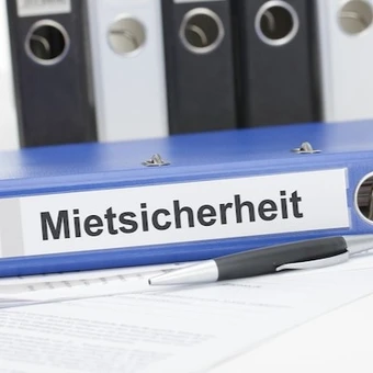 Blauer Aktenordner mit der Aufschrift „Mietsicherheit“, daneben ein Kugelschreiber und Dokumente – Symbolbild für Verwaltung und Absicherung von Mietverhältnissen durch eine Mietkautionsversicherung.