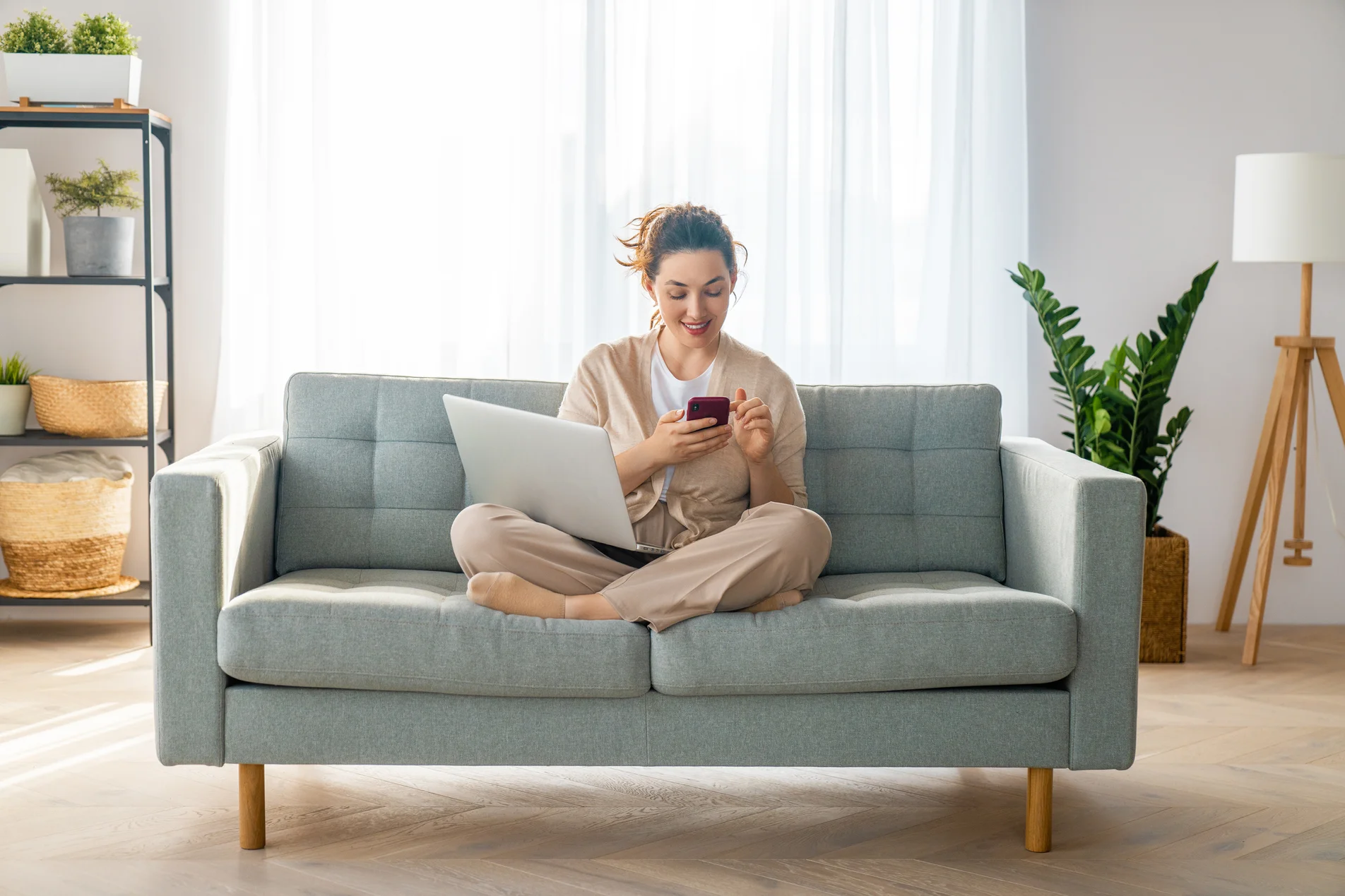 Junge Frau sitzt entspannt auf dem Sofa mit Laptop und Smartphone – Symbolbild für digitalen Abschluss einer Mietkautionsversicherung von zu Hause aus.