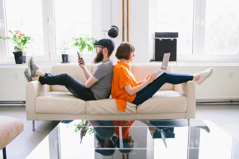 Junges Paar sitzt Rücken an Rücken auf Sofa, nutzt Laptop und Smartphone – Symbolbild für digitale Antragstellung bei einer Mietkautionsversicherung.