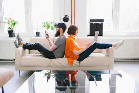 Junges Paar sitzt Rücken an Rücken auf Sofa, nutzt Laptop und Smartphone – Symbolbild für digitale Antragstellung bei einer Mietkautionsversicherung.